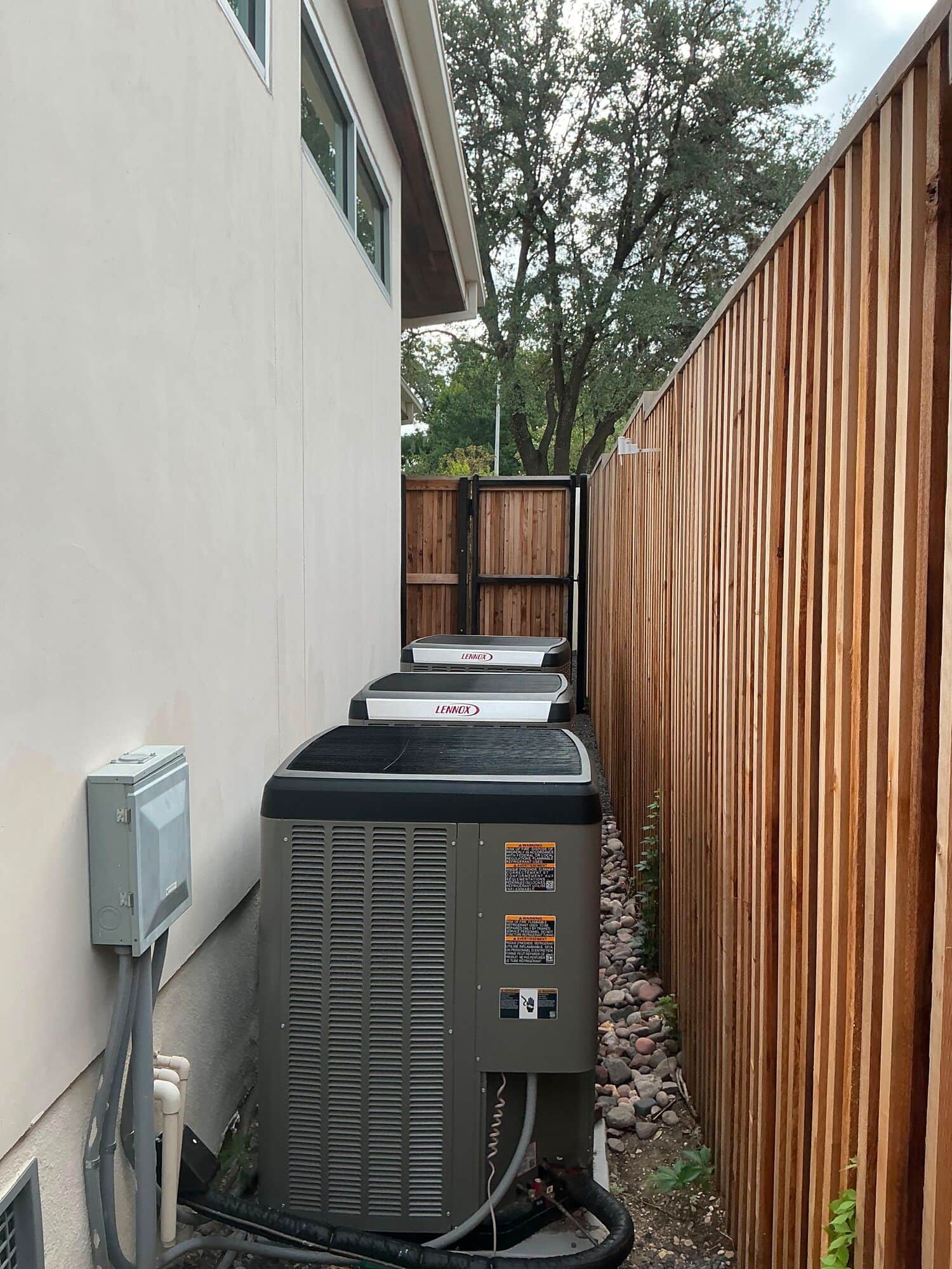 Three Lennox condensing units lined up down the side yard of a modern stucco house, between a new cedar fence and a river-rock bed, with the electrical disconnect on the wall.