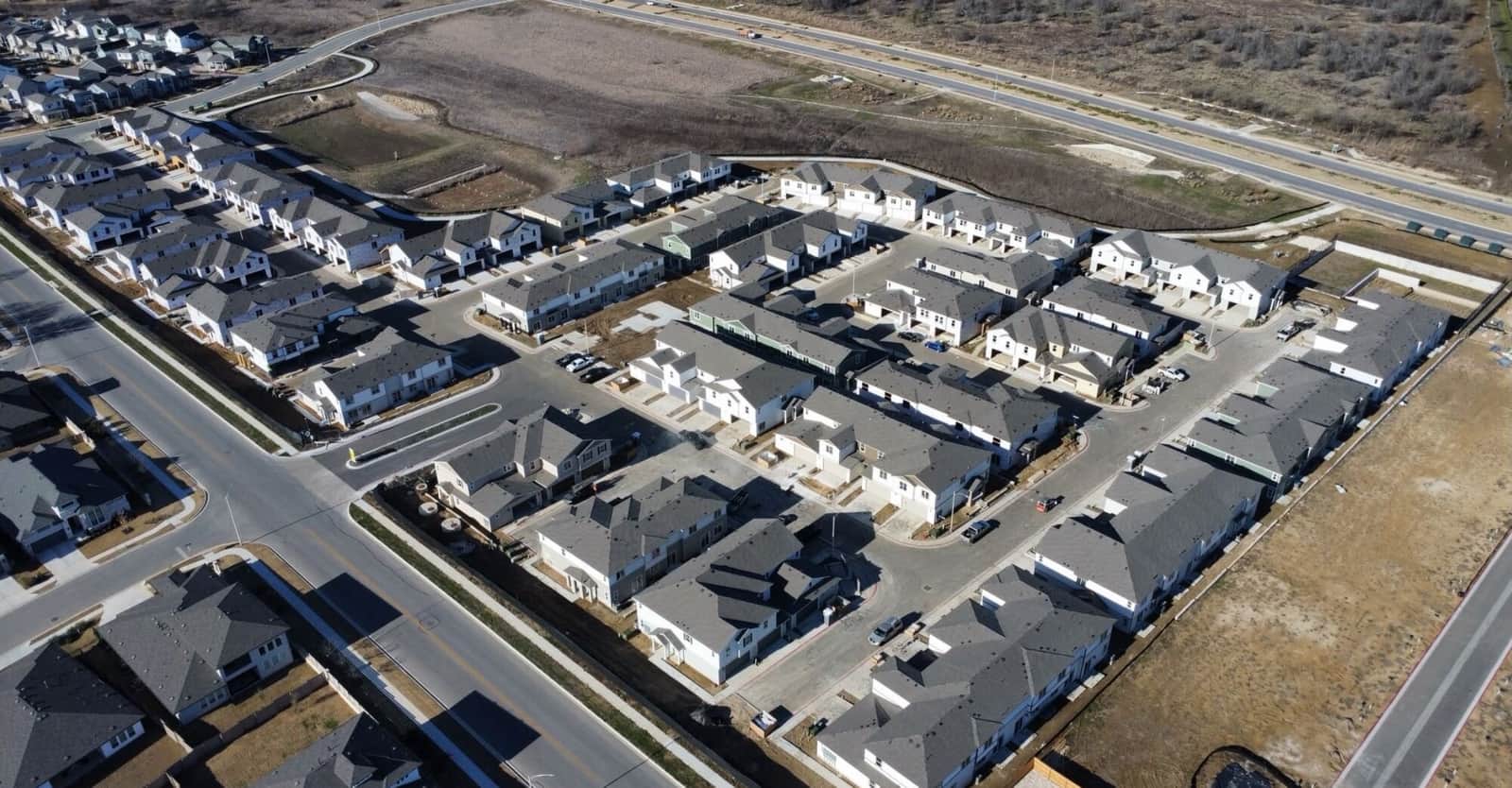 Aerial view over a new Central Texas subdivision — rows of finished and part-built houses on fresh streets, with open field and a highway beyond.