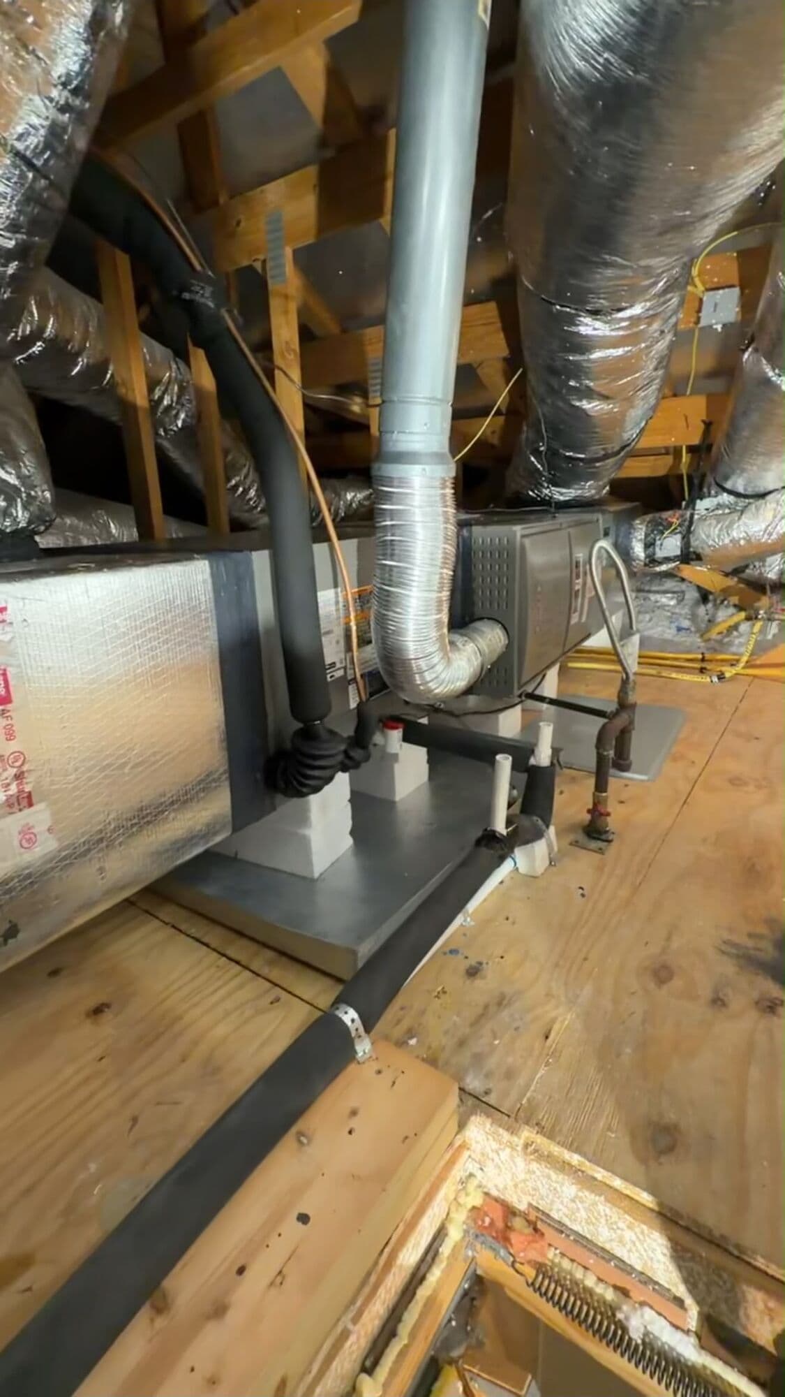 Attic air handler and insulated supply plenum on a plywood platform, with a flexible flue connector, PVC condensate lines and an insulated refrigerant line set running out across the decking.