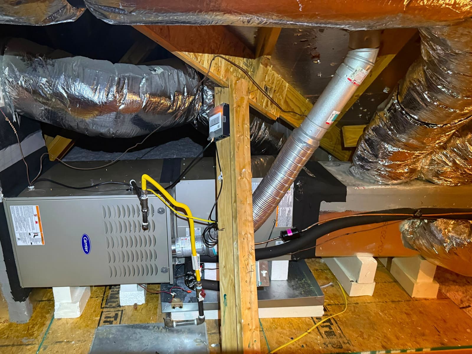 Carrier gas furnace standing on an attic platform, with a yellow flexible gas connector, a metal flue rising through the framing and insulated flex duct runs overhead.