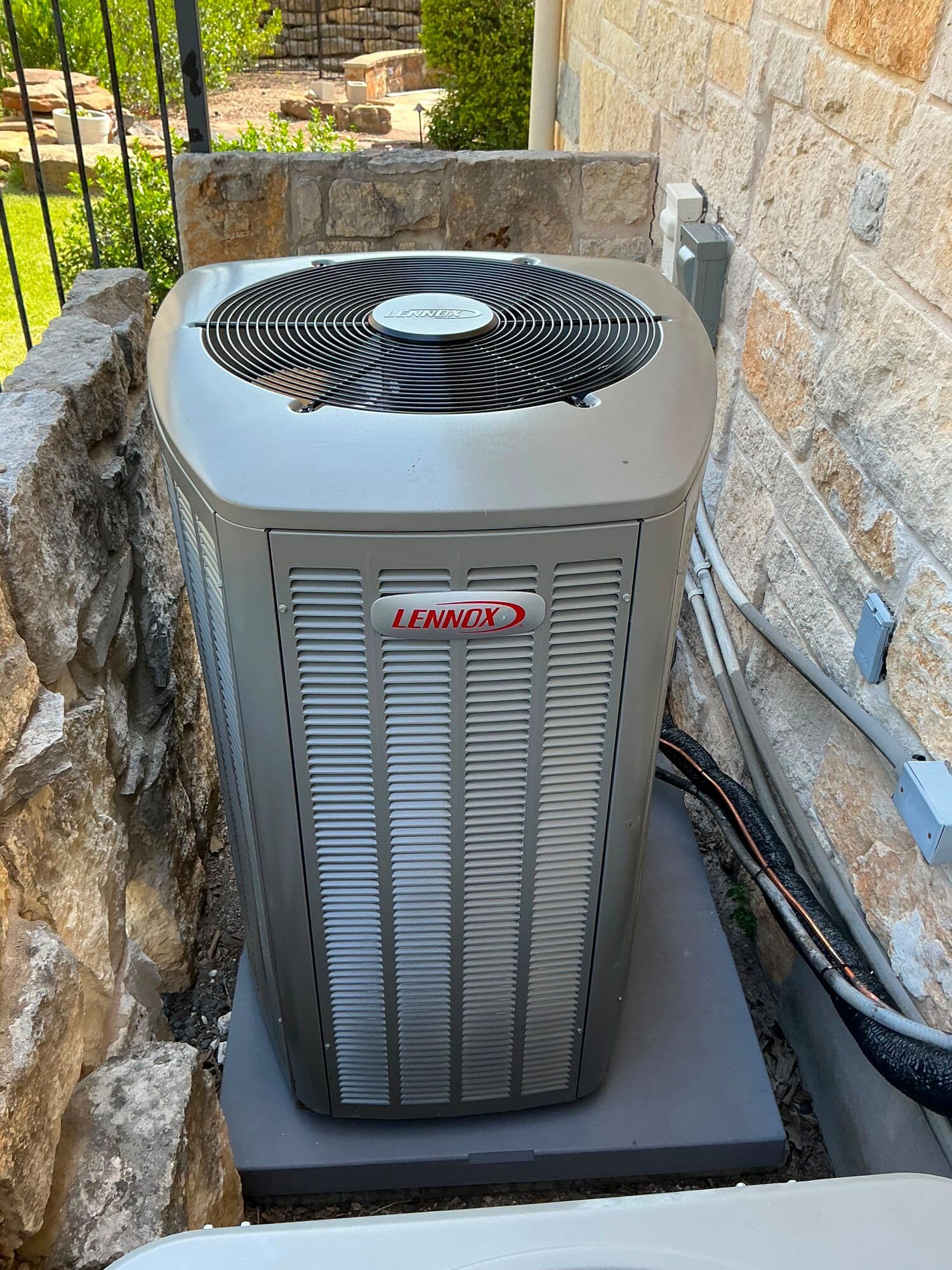 Lennox condensing unit on a pad against a Texas limestone wall, its refrigerant line set and electrical disconnect run up the stone, with an iron fence and green lawn behind it.