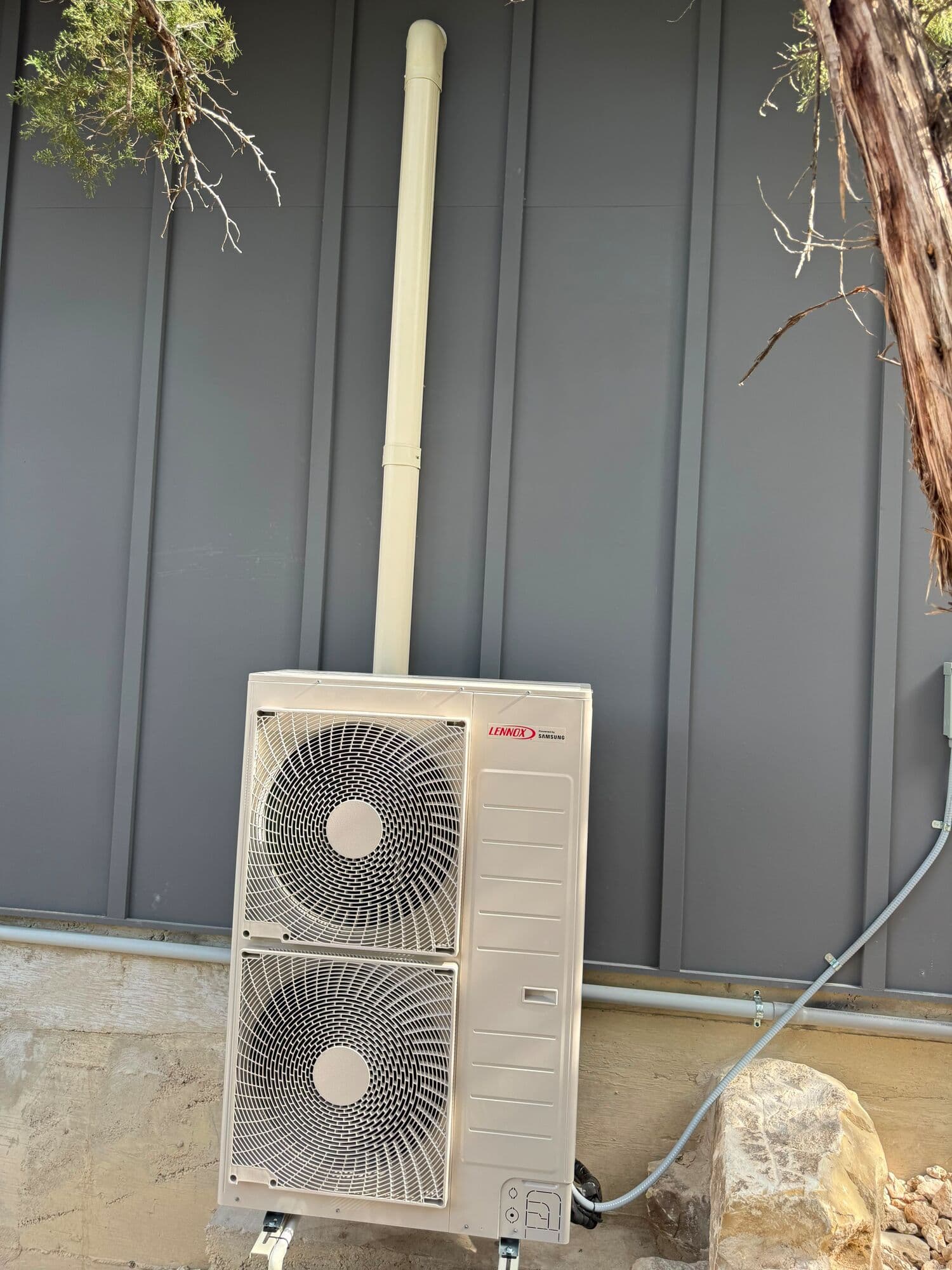 Two-fan Lennox mini-split condensing unit mounted against a grey metal-panel wall, its line-set chase running straight up the panel to the roof and a disconnect box beside it.