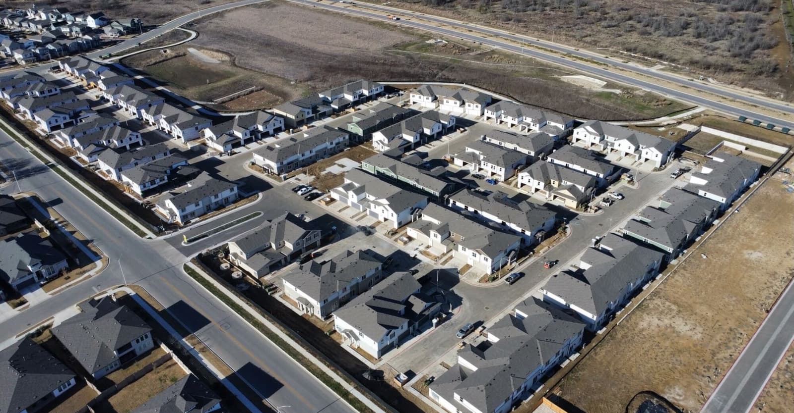 Aerial view over a new Central Texas subdivision — rows of finished and part-built houses on fresh streets, with open field and a highway beyond.